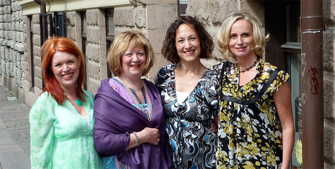 Fyra kvinnor bredvid varandra, en har sljut gröna klder, en har lila kläder och de andra två mönstrade kläder